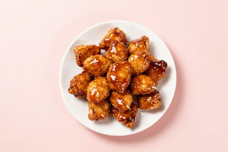 Glossy honey-BBQ chicken bites piled on a white ceramic plate with toothpicks against a pale pink backdrop.