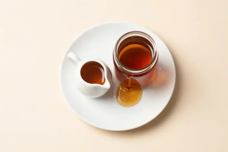 Glossy amber-brown carrot cake syrup in a white pitcher beside a full glass jar