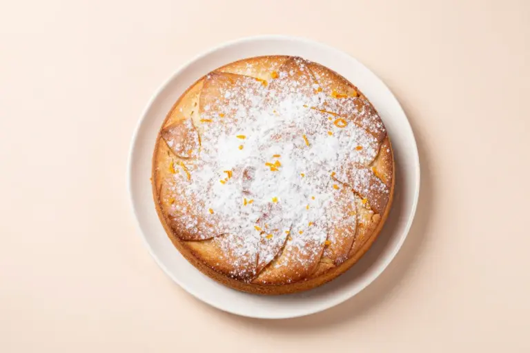 Sicilian whole orange cake on a white plate, dusted with confectioners' sugar against a pastel-blush backdrop.