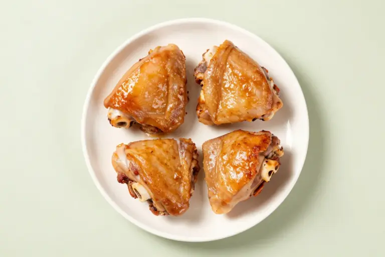 Four bone-in, skin-on chicken thighs with deeply browned skin on a white ceramic plate.